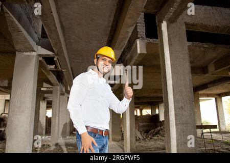 Porträt eines glücklichen jungen, gutaussehenden indischen Bauingenieurs oder Architekten, der ein weißes Hemd und einen gelben Schutzhelm trägt und beim Konstrukt Daumen nach oben zeigt Stockfoto