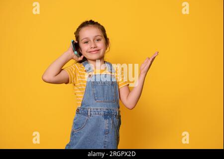 Süßes kleines Mädchen spricht mit dem Handy, lächelt, schönes, zahniges Lächeln schaut in die Kamera, trägt ein gelb gestreiftes T-Shirt und ein Jeanskleid, isoliert Stockfoto