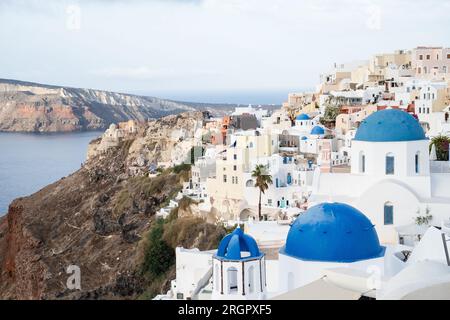 Fotografieren Sie die berühmten Kirchen mit blauen Kuppeln in Oia, Santorin, inmitten von Gebäuden an den Klippen, mit einem wolkigen Himmel darüber. Stockfoto