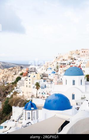 Fotografieren Sie die berühmten Kirchen mit blauen Kuppeln in Oia, Santorin, inmitten von Gebäuden an den Klippen, mit einem wolkigen Himmel darüber. Stockfoto