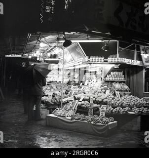 1950er, historisch, ein nasser Abend und ein Mann mit Schirm an einem Straßenstand, der Obst in Tokio, Japan, verkauft. Stockfoto