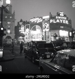 1960er Uhr, historisch, Abendzeit am Londoner West End, mit Piccadilly Circus und seinen berühmten Neon-Werbeschildern beleuchtet. Autos der Epoche, einschließlich eines Londoner Taxis an der Ampel, bereit für den berühmten Kreisverkehr, der 1819 gebaut wurde, um Piccadilly mit Regent St. zu verbinden Stockfoto