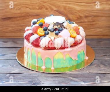 Farbenfroher Kuchen mit Erdbeeren in geschmolzener weißer Schokolade, mit rosafarbener Glasur, frischen Himbeeren, Heidelbeeren und Kumquat. Rustikale Holzmöbel Stockfoto