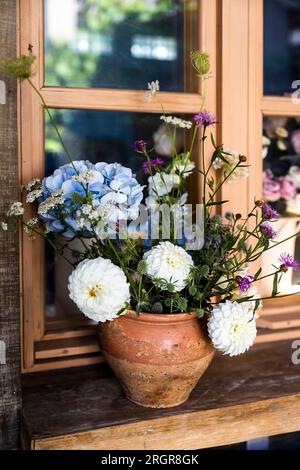Ein rustikaler Strauß aus blauen Hortensien, weißen Chrysanthemen und Dahlien sowie Zichorien in einer braunen Flasche auf einem Holztisch als Dekoration der Veranda Stockfoto