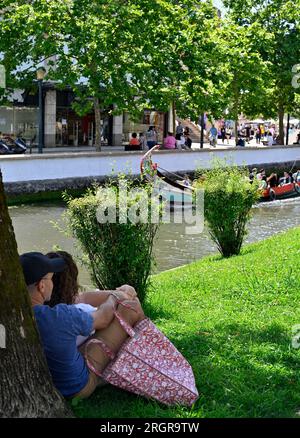 Ein Paar entspannt auf Grasbänken im Schatten sitzen und den traditionellen Kanal Moliceiro Touristenboot Pass im Zentrum von Aveiro, Portugal beobachten Stockfoto