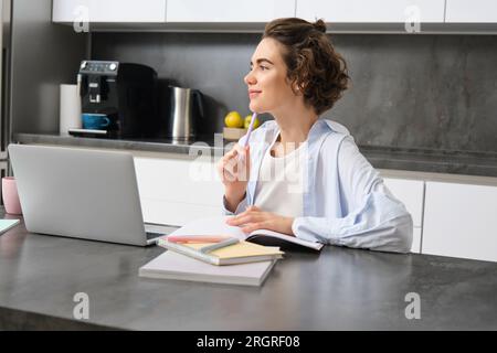 Porträt einer jungen Frau, die denkt, zu Hause an einem Laptop arbeitet, sich Notizen macht, während sie ihre Hausaufgaben macht, Online-Kurse studiert, zuhört Stockfoto