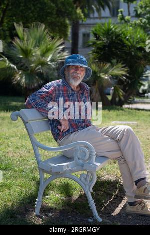 Ein alter Mann mit einem Bart in einem panamahut und einem karierten Hemd sitzt an einem sonnigen Sommertag auf einer Parkbank. Stockfoto