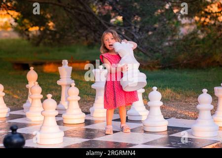 Kleines Weißes Mädchen in einem rosa Kleid, das Straßenschach spielt. An einem Sommerabend spielt ein Kind im Park mit großen Schachfiguren. Stockfoto