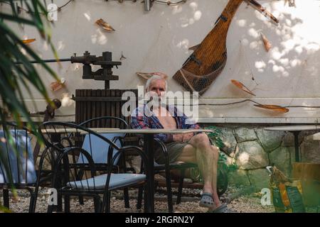 Ein alter Mann in einem karierten Hemd sitzt an einem Tisch, umgeben von Angelmöglichkeiten und Weingütern. Ein alter Bauer und Fischer ruht in seiner eigenen Villa. Stockfoto