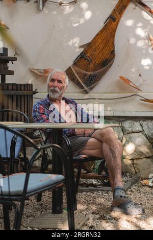 Ein alter Mann in einem karierten Hemd sitzt an einem Tisch, umgeben von Angelmöglichkeiten und Weingütern. Ein alter Bauer und Fischer ruht in seiner eigenen Villa. Stockfoto