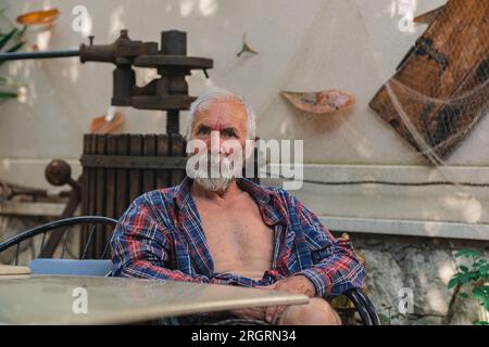 Ein alter Mann in einem karierten Hemd sitzt an einem Tisch, umgeben von Angelmöglichkeiten und Weingütern. Ein alter Bauer und Fischer ruht in seiner eigenen Villa. Stockfoto