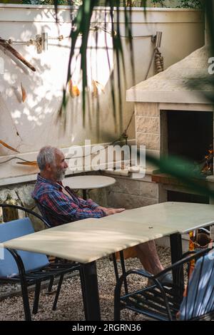 Ein alter Mann in einem karierten Hemd sitzt an einem Tisch, umgeben von Angelmöglichkeiten und Weingütern. Ein alter Bauer und Fischer ruht in seiner eigenen Villa. Stockfoto