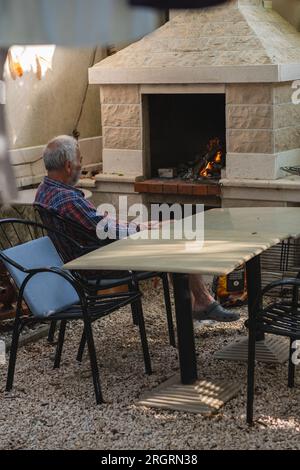 Ein alter Mann in einem karierten Hemd sitzt an einem Tisch, umgeben von Angelmöglichkeiten und Weingütern. Ein alter Bauer und Fischer ruht in seiner eigenen Villa. Stockfoto