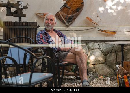 Ein alter Mann in einem karierten Hemd sitzt an einem Tisch, umgeben von Angelmöglichkeiten und Weingütern. Ein alter Bauer und Fischer ruht in seiner eigenen Villa. Stockfoto
