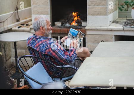 Ein alter Mann in einem karierten Hemd sitzt am Tisch und liest Zeitung. Ein alter Bauer und Fischer ruht in seiner eigenen Villa. Stockfoto