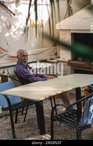 Ein alter Mann in einem karierten Hemd sitzt an einem Tisch, umgeben von Angelmöglichkeiten und Weingütern. Ein alter Bauer und Fischer ruht in seiner eigenen Villa. Stockfoto