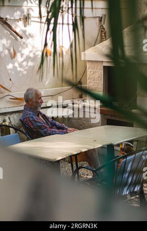 Ein alter Mann in einem karierten Hemd sitzt an einem Tisch, umgeben von Angelmöglichkeiten und Weingütern. Ein alter Bauer und Fischer ruht in seiner eigenen Villa. Stockfoto