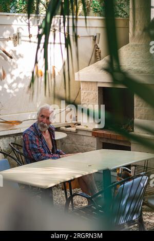 Ein alter Mann in einem karierten Hemd sitzt an einem Tisch, umgeben von Angelmöglichkeiten und Weingütern. Ein alter Bauer und Fischer ruht in seiner eigenen Villa. Stockfoto