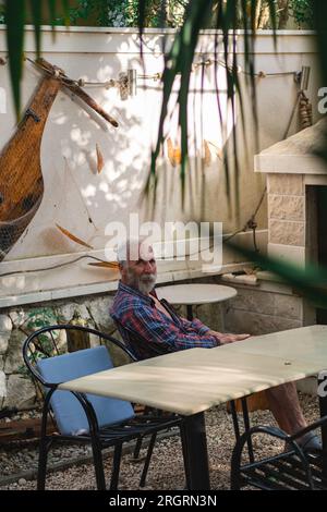 Ein alter Mann in einem karierten Hemd sitzt an einem Tisch, umgeben von Angelmöglichkeiten und Weingütern. Ein alter Bauer und Fischer ruht in seiner eigenen Villa. Stockfoto