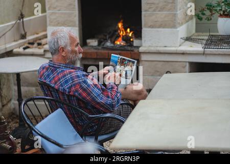 Ein alter Mann in einem karierten Hemd sitzt am Tisch und liest Zeitung. Ein alter Bauer und Fischer ruht in seiner eigenen Villa. Stockfoto