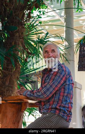 Ein alter Mann in einem karierten Hemd sitzt an einem Tisch, umgeben von Angelmöglichkeiten und Weingütern. Ein alter Bauer und Fischer ruht in seiner eigenen Villa. Stockfoto