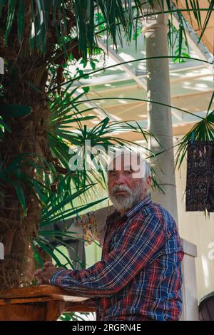 Ein alter Mann in einem karierten Hemd sitzt an einem Tisch, umgeben von Angelmöglichkeiten und Weingütern. Ein alter Bauer und Fischer ruht in seiner eigenen Villa. Stockfoto