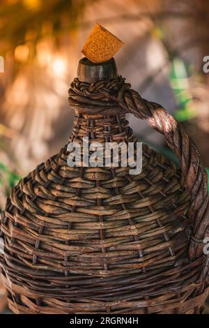 Große Weinflasche In Wicker-Umhüllung Im Vintage-Stil. Der alte Demijohn mit Kork in den Sonnenstrahlen auf einem Glastisch mit Weinkorken unter Glas. Stockfoto