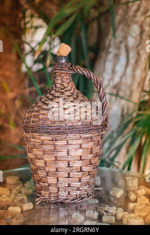 Große Weinflasche In Wicker-Umhüllung Im Vintage-Stil. Der alte Demijohn mit Kork in den Sonnenstrahlen auf einem Glastisch mit Weinkorken unter Glas. Stockfoto