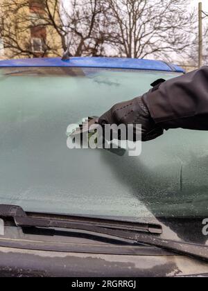 Autofenster im Winter reinigen. Der Mensch kratzt mit einem Plastikkratzer von der Windschutzscheibe eines blauen Autos. Hand in schwarzem Lederhandschuh im C Stockfoto