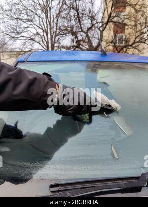 Autofenster im Winter reinigen. Der Mensch kratzt mit einem Plastikkratzer von der Windschutzscheibe eines blauen Autos. Hand in schwarzem Lederhandschuh im C Stockfoto