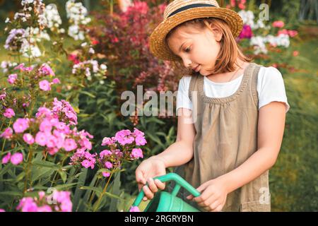 Bauernmädchen im Sommerhut. Ein kleiner Gärtner, der Blumenbeet mit rosa Blumen bewässert, Spaß im Garten hat. Große grüne Gießkanne Wasser. Ha Stockfoto