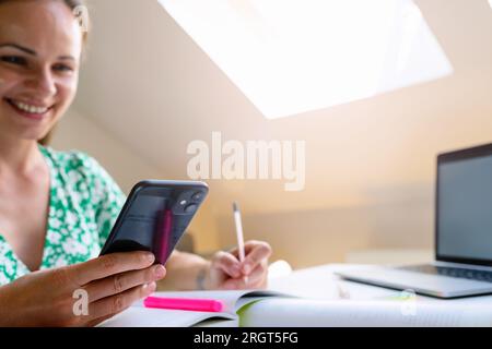 Junge brünette Frau, die ihr Handy lächelt und sich Notizen in ihr Notizbuch schreibt, während sie zu Hause am Schreibtisch sitzt. Stockfoto