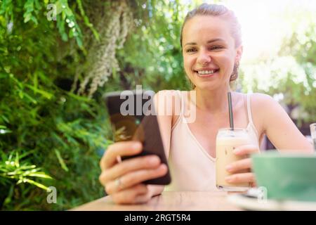 Eine junge Frau aus der Stadt nutzt ihr Smartphone und lächelt, während sie im Sommercafé im Freien Eiskaffee trinkt. Stockfoto