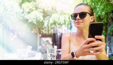 Eine junge Frau in der Stadt, die eine Sonnenbrille trägt, sitzt im Sommercafé im Freien am Tisch und hält ihr Smartphone in den Händen und lächelt. Stockfoto