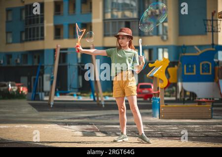 Ein süßes Mädchen im Schulalter bläst im Hof eines mehrstöckigen Gebäudes im Sommer große Seifenblasen. Sommerkonzept, Spiele im Freien Stockfoto