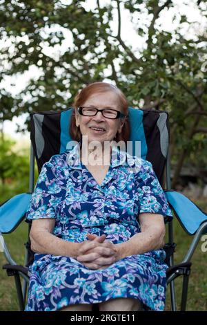 Porträt einer glücklichen lächelnden Großmutter im Garten zu Hause. Alte Frau ruht sich im Stuhl aus, schaut in die Kamera, Obstgarten im Hintergrund. Summe Stockfoto
