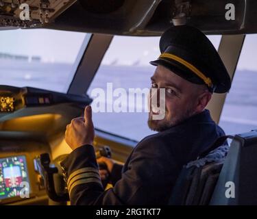 Kaukasischer bärtiger Mann, der beim Fahren eines Flugsimulators lächelt. Der Pilot im Cockpit zeigt Daumen nach oben. Stockfoto