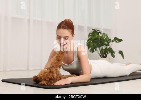 Glückliche junge Frau, die Yoga auf Matte mit ihrem süßen Hund drinnen praktiziert Stockfoto