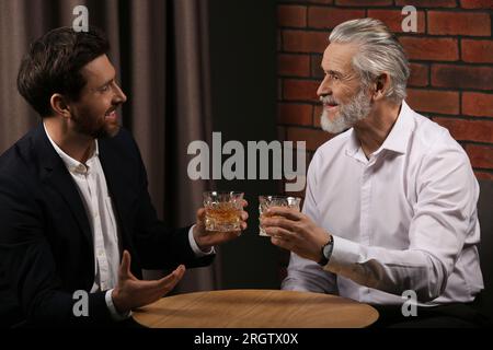 Männer mit Whiskey-Gläsern, die sich drinnen an einem Holztisch unterhalten Stockfoto