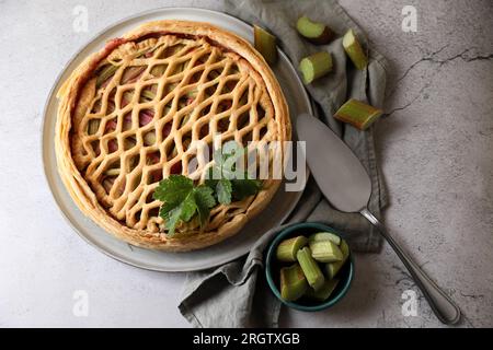 Frisch gebackener Rhabarberkuchen, geschnittene Stängel und Kuchenservice auf hellgrauem Tisch, flach liegend Stockfoto