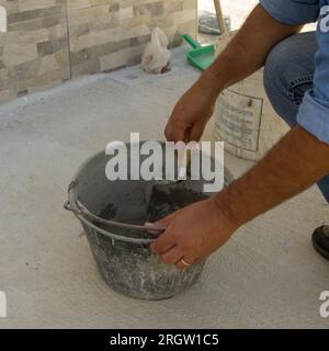 Bild einer Maurerhände, die Zement und Kleber in einem Eimer mischt. Manuelle Mauerwerk- und Fliesenarbeiten. Stockfoto