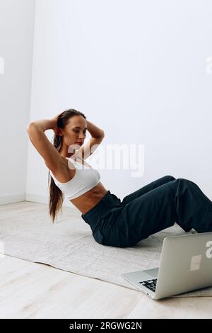 Eine Frau geht auf dem Boden Sport treiben, mit einem Online-Trainer über einen Laptop. Sport in Abriegelung Stockfoto