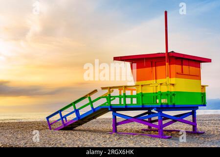 Der mit einem Regenbogen aus Farbtönen geschmückte Rettungsschwimmturm steht hoch vor der malerischen Kulisse des Ozeans und einem atemberaubenden Sonnenuntergang. Küstenatmosphäre und Stockfoto