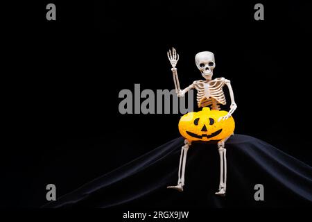 Ein menschliches Skelett, das winkt, einen orangefarbenen Halloween-Kürbis auf schwarzem Hintergrund hält. Halloween-Grußkarte Stockfoto