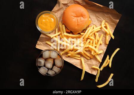 Cheeseburger und Pommes frites auf braunem Papier und Holztisch, Draufsicht. Burger, Pommes Frites, Cola mit Eis in einem Glas und Senf. Fastfood Stockfoto