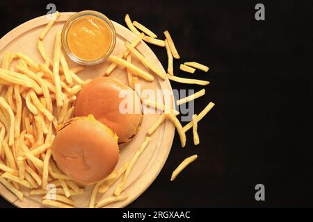 Cheeseburger und Pommes Frites auf einem Schneidebrett, Draufsicht, dunkler Holzhintergrund. Fastfood. Burger, Pommes frites und Senf Stockfoto