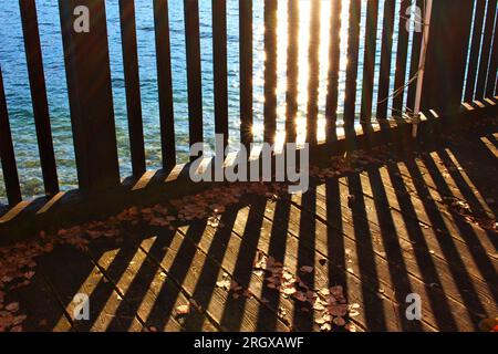 Die Sonne reflektiert über dem See und scheint auf einen Holzzaun und wirft Schatten auf die Holzterrasse. Cooler, symmetrischer Hintergrund am Sommerabend Stockfoto