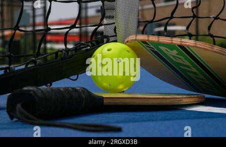 West Islip, New York, USA - 17. Juli 2023: Zwei Pickleball-Paddel und ein gelber Peitschenball dazwischen auf blauem Platz aus nächster Nähe. Stockfoto