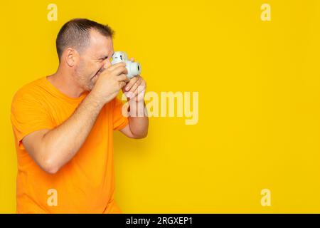 Glücklicher hispanischer Mann in einem T-Shirt, der lächelt und ein Profilbild mit einer kleinen Fotokamera vor gelbem Hintergrund macht. Stockfoto