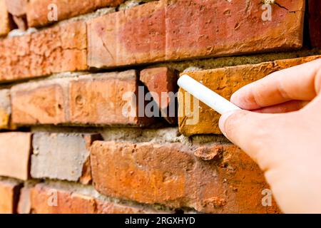 Halten Sie weiße Schulkreide in der Hand, bereit zum Schreiben oder Zeichnen auf roten Ziegeln einer veralteten verfallenen beschädigten Wand. Stockfoto